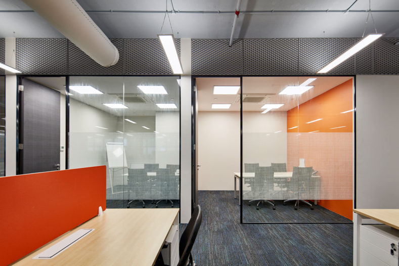 Acromec Ltd Offices. Director's offices align to the core wall. The overall aesthetic is industrial, with an exposed soffit, fabric ducts, cable trays and trunking. Expanded mesh panels above the rooms screen the acoustic baffles.